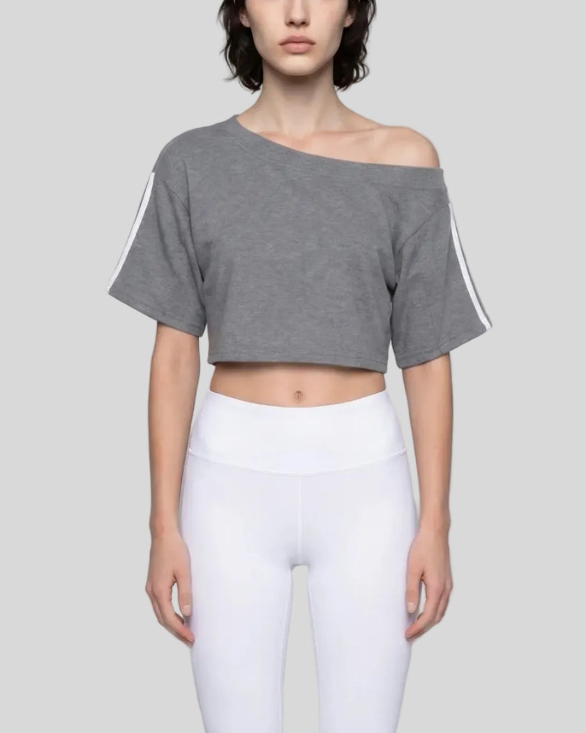 Mid body front view of the Athlesi TrackLine Off-Shoulder Tee in gray styled in a minimalist architectural setting