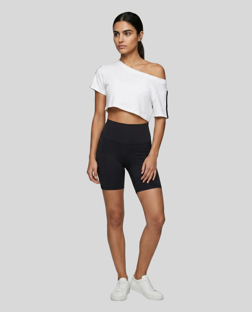 Full body front view of a woman wearing the Athlesi TrackLine Off-Shoulder Tee in white styled for modern athleisure movement