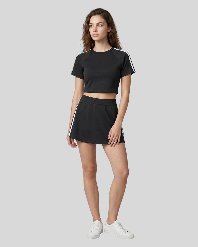 Full body front view of a woman wearing the Athlesi TrackLine Performance Skort in black styled with the TrackLine Performance Crop Tee