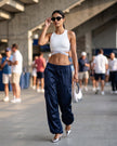 A woman walking in a stadium-like corridor wearing a white TrackLine Rally Sports Bra with a loose dark navy wide-leg cargo pants, and metallic silver pointed shoes. She is holding a silver handbag and wearing dark sunglasses.