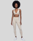 A woman modeling a matching light apricot TrackLine Riviera activewear set consisting of a sports bra with white piping and high-waisted leggings with white side stripes, paired with white sneakers, against a plain white studio background.