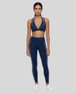 A woman modeling a matching navy blue TrackLine Riviera activewear set consisting of a sports bra with white piping and high-waisted leggings with white side stripes, paired with white sneakers, against a plain white studio background.