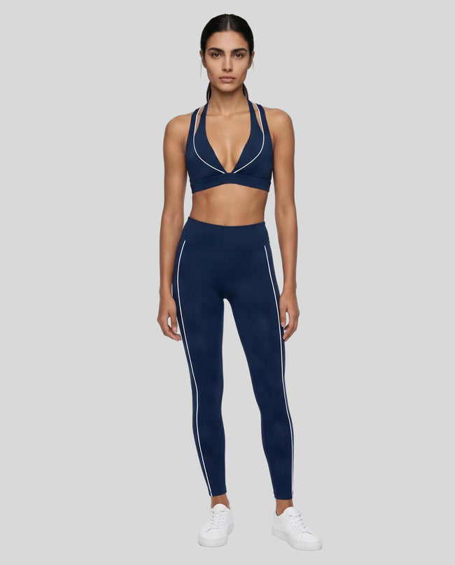 A woman modeling a matching navy blue TrackLine Riviera activewear set consisting of a sports bra with white piping and high-waisted leggings with white side stripes, paired with white sneakers, against a plain white studio background.