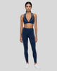 A woman modeling a matching navy blue TrackLine Riviera activewear set consisting of a sports bra with white piping and high-waisted leggings with white side stripes, paired with white sneakers, against a plain white studio background.