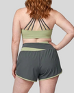 Back view of a woman modeling a dark gray and green bean TrackLine Synergy Performance Shorts with a matching sports bra.