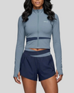 A woman modeling TrackLine Synergy Perfomance Shorts in navy blue with gray blue contrast details on the waist, paired with TrackLine Synergy Perfomance Jacket with a front zipper, horizontal navy stripe detail, and a small white 'Athlesi' logo on the chest.