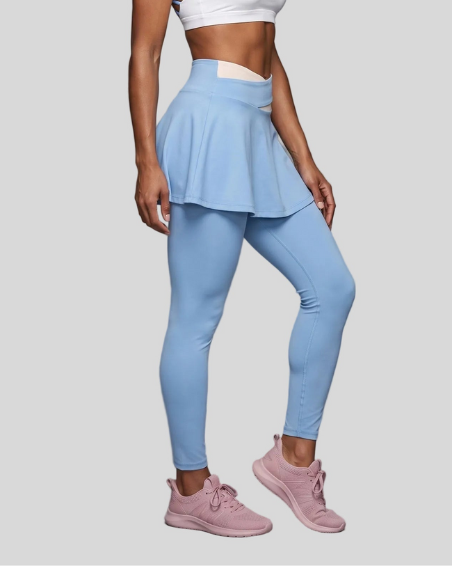 A model wearing light blue TrackLine Synergy Skirted Leggings with a flared pleated overlay and a apricot waistband, paired with a white sports bra and pink sneakers.