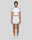 Woman wearing the Athlesi TrackLine Zip Crop Tee in white styled with Athlesi TrackLine Slit Skirt, in a modern minimalist setting