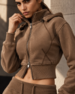 A woman wearingAthlesi Vintage Washed Edition Hoodie in a warm taupe-brown color, paired with matching drawstring pants, photographed outdoors against a stone wall.
