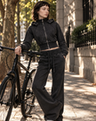 A woman stands on a city sidewalk beside a black bicycle, wearing a black Athlesi Vintage Washed Edition hoodie and matching wide-leg pants. The setting includes trees, stone buildings, and a black railing.